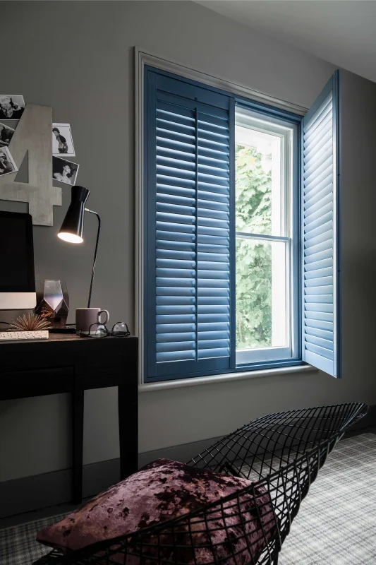 Blue full-height plantation shutters fitted to a bedroom window beside a desk and hammock.