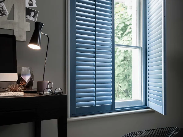 Blue full-height plantation shutters fitted to a bedroom window beside a desk and hammock.