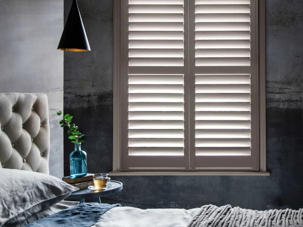 Cockleshell plantation shutters fitted to a bedroom window, providing elegant light control, privacy and a warm neutral finish.