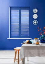 Lindos blue plantation shutters fitted to a dining room window with matching blue walls.