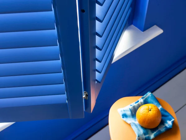 Lindos blue plantation shutter panel opened beside a small table in a vibrant blue interior.