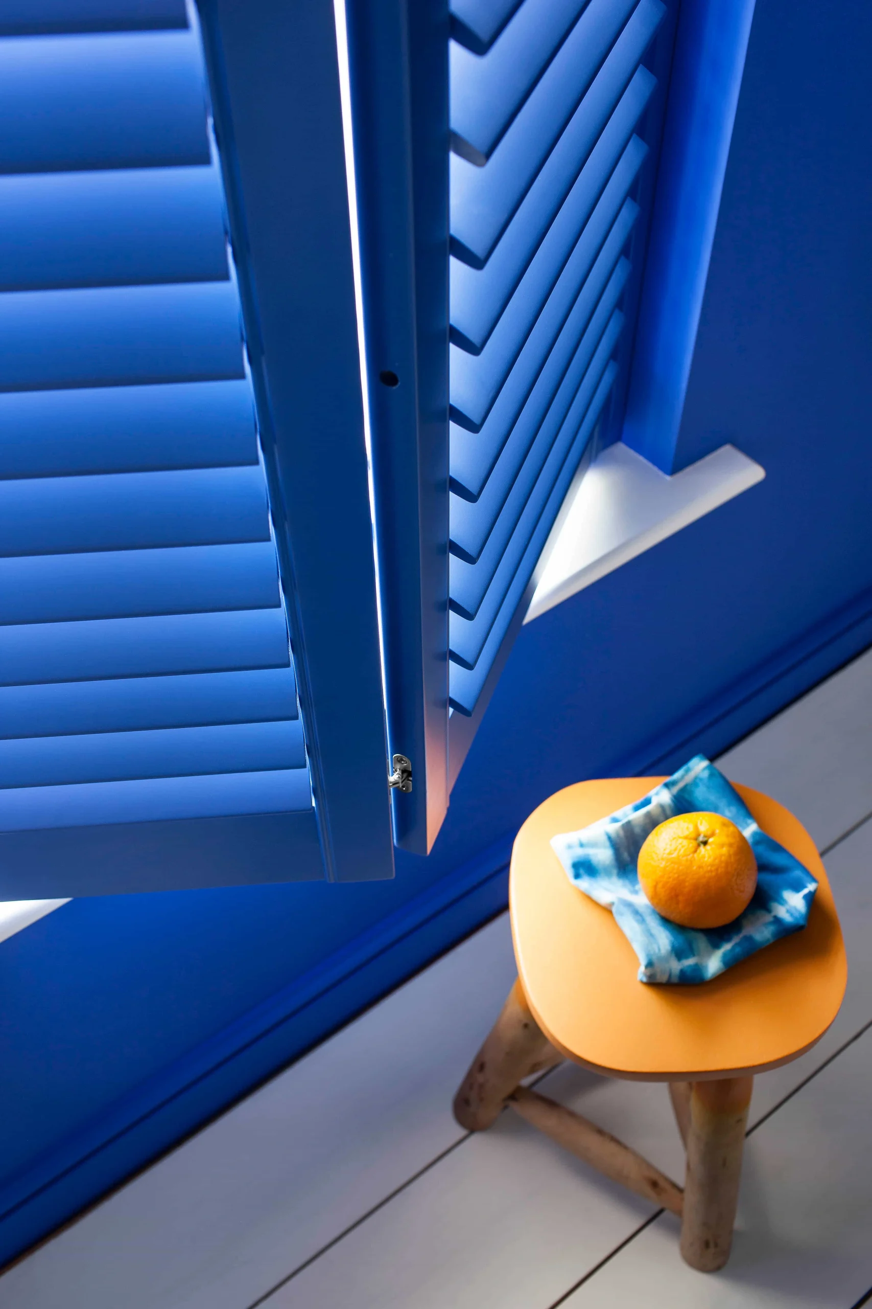 Lindos blue plantation shutter panel opened beside a small table in a vibrant blue interior.