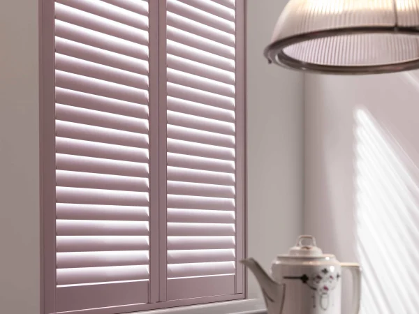 Pastel pink plantation shutters filtering sunlight beside a dining table.