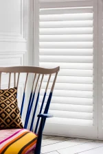 White full-height plantation shutters fitted beside a colourful accent chair in a bright living room.