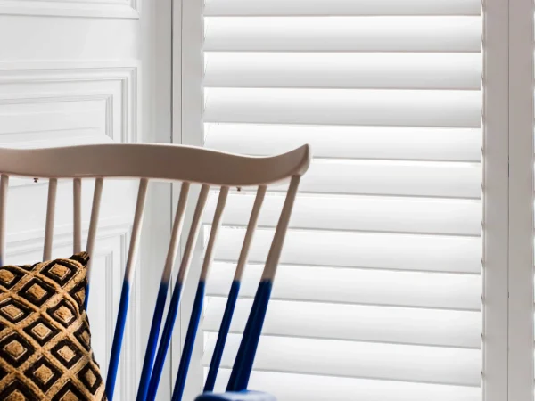 White full-height plantation shutters fitted beside a colourful accent chair in a bright living room.