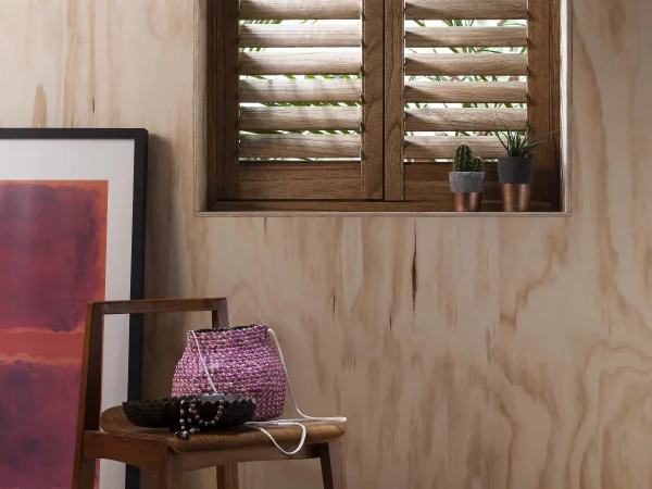 Wood-grain plantation shutters fitted to a small window in a natural plywood-lined room.
