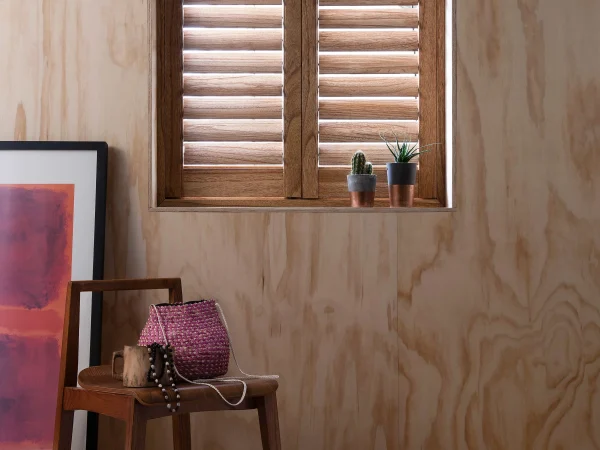 Wood-grain plantation shutters closed across a small window above a side chair.